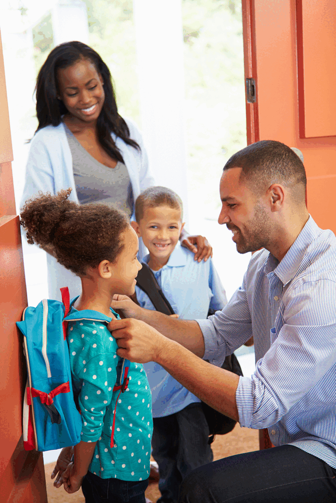 a father finishes putting a backpack on his child as a smiling mother and sibling wait